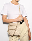 MZ Wallace Downtown Crosby Crossbody Buff Crossbody worn by model wearing a white T-Shirt and beige trousers in front of white background.