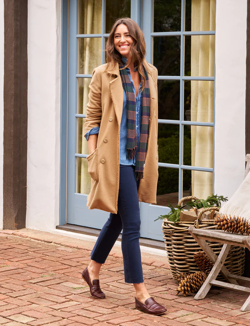 Frank & Eileen Sweatshirt Trench Coat Camel on a brunette model also wearing a  denim shirt, navy jeans, a plaid scarf and brown loafers walking on a brick path in front of windows outside