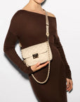 MZ Wallace Small Crosby Lock Crossbody Buff Crossbody worn by model wearing brown off the shoulder dress in front of white background. 