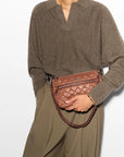 model wearing a brown sweater and green trousers holding  the mz wallace Crosby Ella Crossbody Terracotta on a grey background