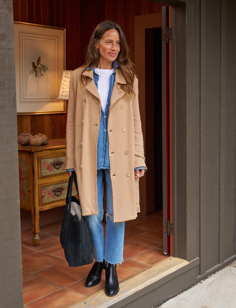 Frank & Eileen Sweatshirt Trench Coat Camel on a brunette model also wearing a white tee, denim shirt, jeans, black boots, and holding a black tote standing in a doorframe of a house