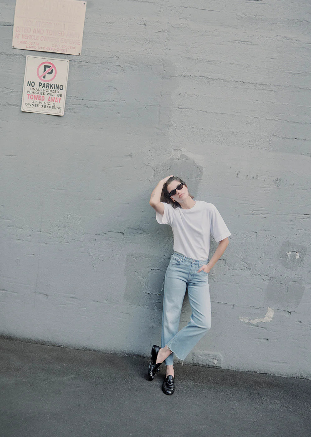 brunette model leaning up against a concrete building outside wearing the citizens of humanity Palma straight Lisbon jeans with black loafers and a white tee 