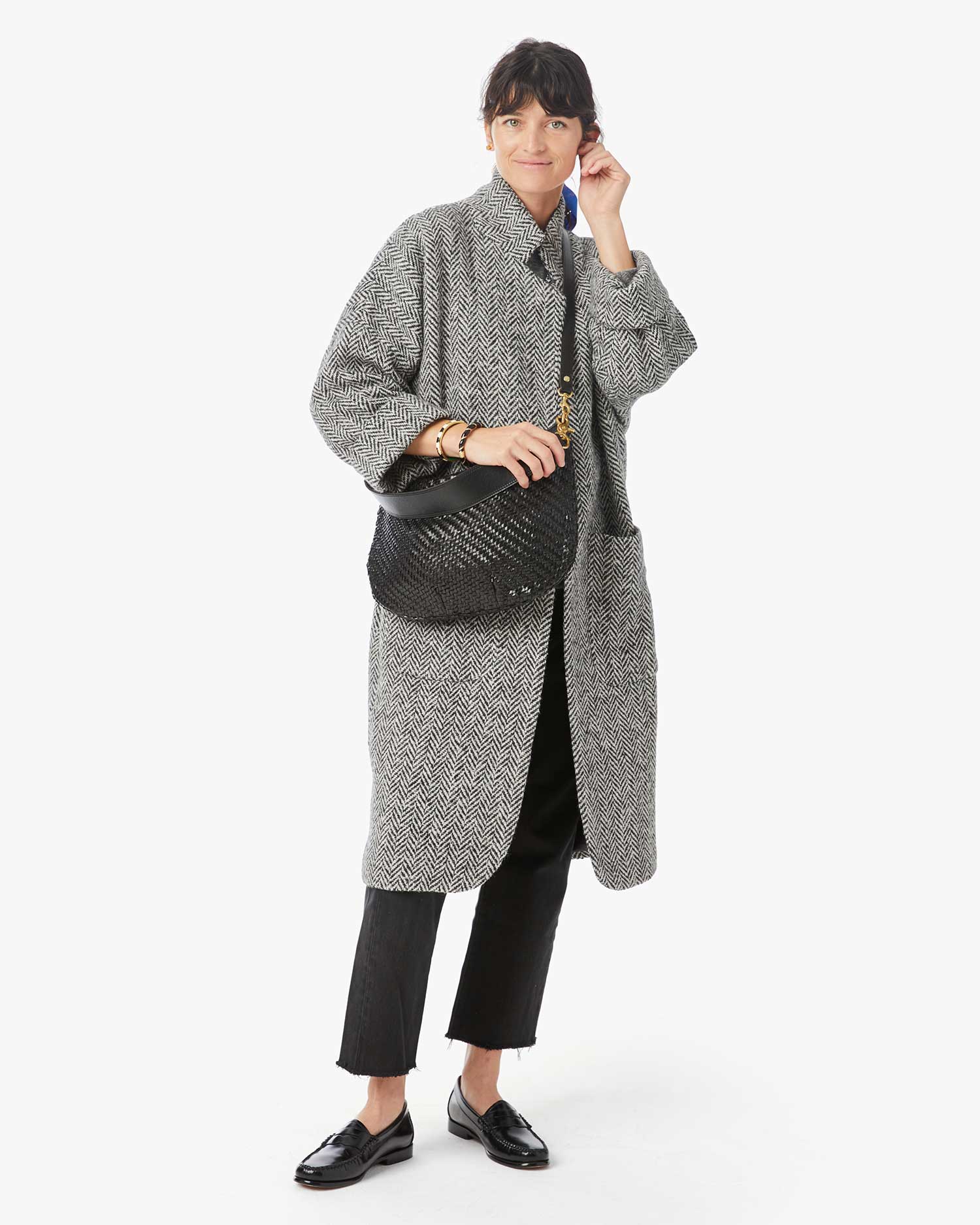 brunette model wearing a grey houndstooth long coat with black jeans and black loafers and wearing the Clare v Moyen Messenger Black Woven Zig Zag on a white background