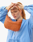 red haired model wearing a blue dress holding the Clare v Petit Moyen Messenger Natural Woven Checker around her face on a white background