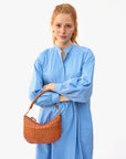 red haired model wearing a blue dress holding the Clare v Petit Moyen Messenger Natural Woven Checker on her arm with her arms crossed on a white background