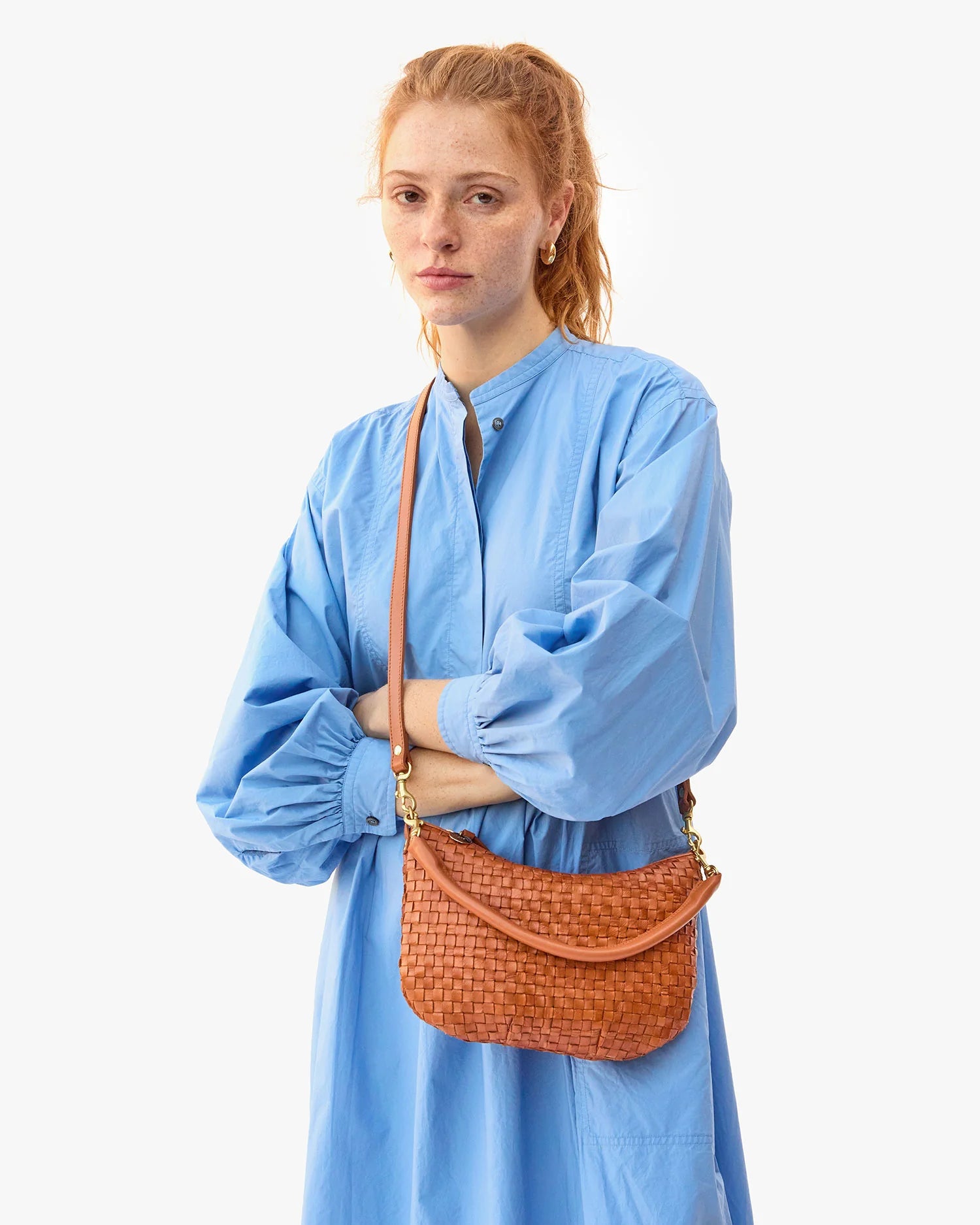 red haired model wearing a blue dress holding the Clare v Petit Moyen Messenger Natural Woven Checker as a crossbody with her arms crossed on a white background