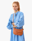 red haired model wearing a blue dress holding the Clare v Petit Moyen Messenger Natural Woven Checker as a crossbody with her arms crossed on a white background