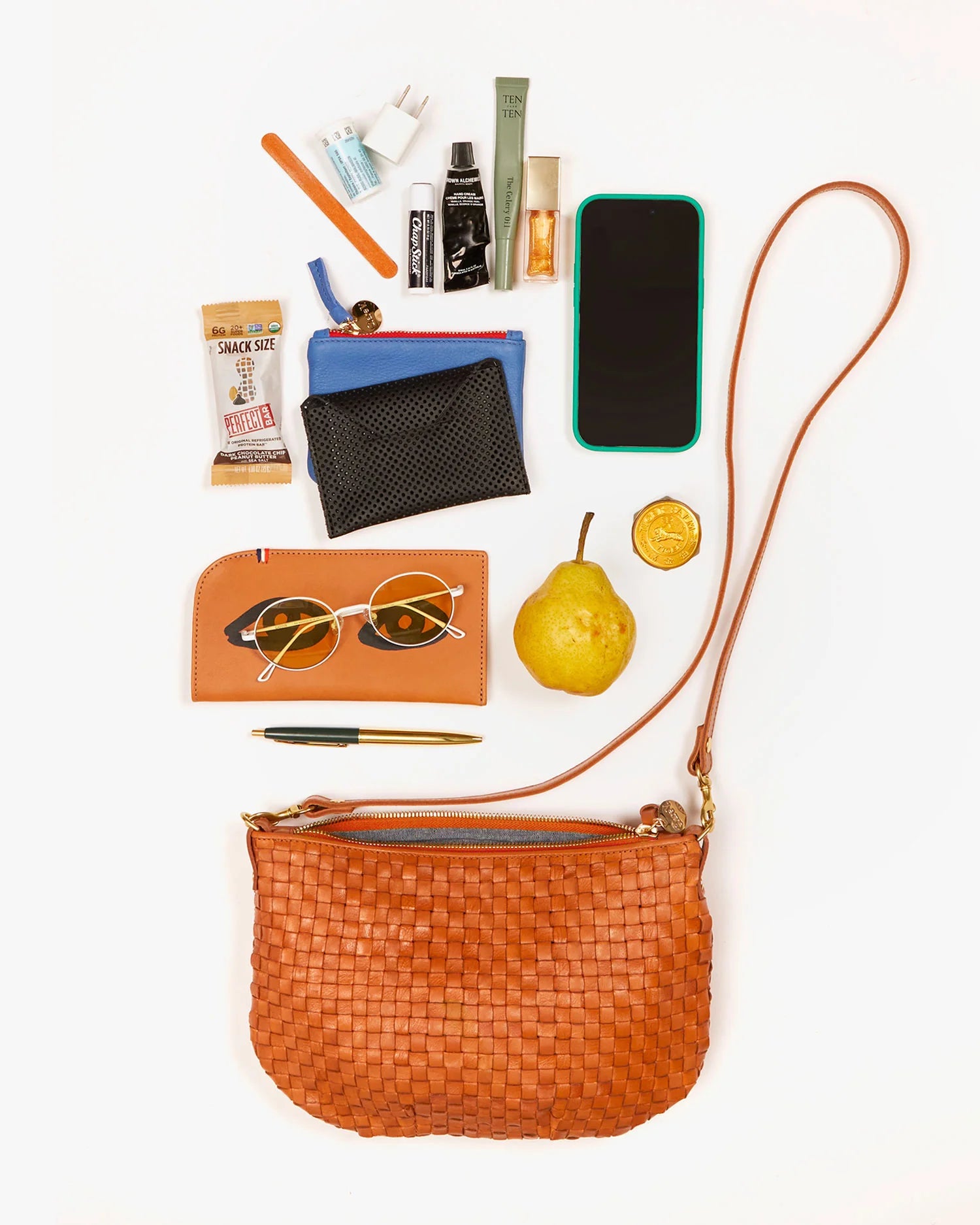 the Clare v petit moyen messenger in natural checker on a black background showing various items that can fit in it like a pear, sunglasses, and a phone