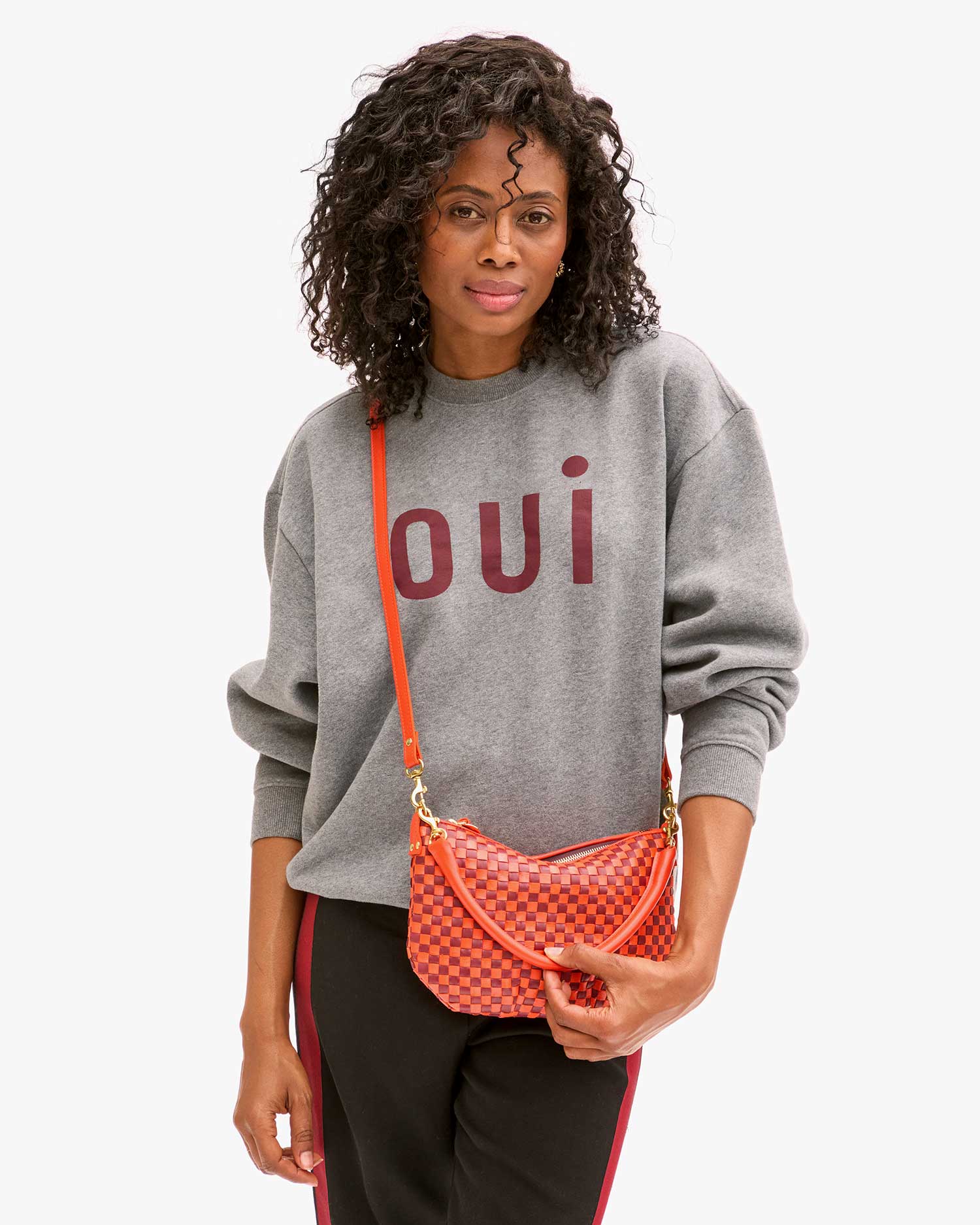 black model wearing a grey Oui sweatshirt and black track pants and wearing the Clare v Petit Moyen Messenger Bright Poppy/Bordeaux Woven Checker on a white background