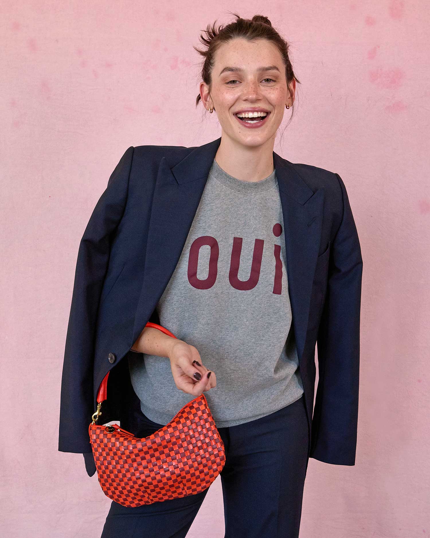 brunette model in front of a pink background wearing a Oui grey t-shirt, navy blazer draped over her shoulders, matching trousers, and holding the Clare v Petit Moyen Messenger Bright Poppy/Bordeaux Woven Checker on her arm while smiling