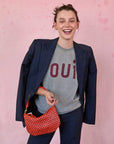 brunette model in front of a pink background wearing a Oui grey t-shirt, navy blazer draped over her shoulders, matching trousers, and holding the Clare v Petit Moyen Messenger Bright Poppy/Bordeaux Woven Checker on her arm while smiling