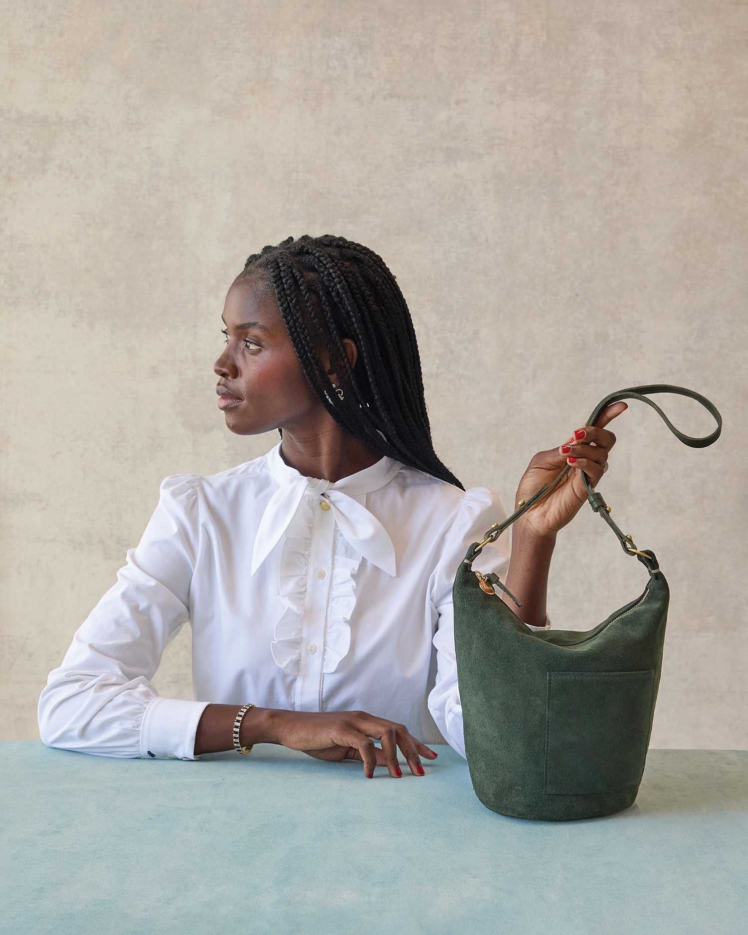 black model wearing a white button up sitting at a blue table in front of a grey background holding the straps of the Clare v petite Jeanne laden suede as it sits on the table