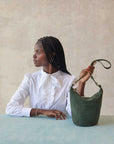 black model wearing a white button up sitting at a blue table in front of a grey background holding the straps of the Clare v petite Jeanne laden suede as it sits on the table