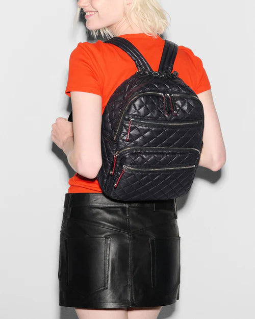 back of a blonde model wearing mz Wallace Small Crosby Backpack Black, black leather skirt, and an orange tee on a white background