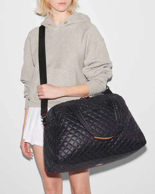 MZ Wallace Travel Jim Black Tote & Satchel, worn by blonde model using the shoulder strap wearing a grey hoodie and white shorts. 
