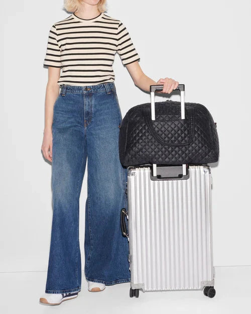 MZ Wallace Travel Jim Black Tote & Satchel sitting on silver luggage held onto by blonde model wearing a stripped short sleeve and blue wide legged pants and sneakers on feet. 
